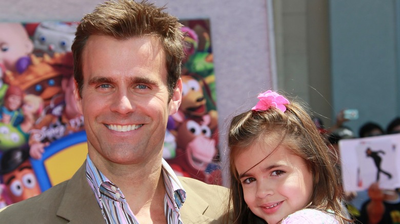 Cameron Mathison wearing a suit, holding Leila Mathison wearing a flower bow on her head.