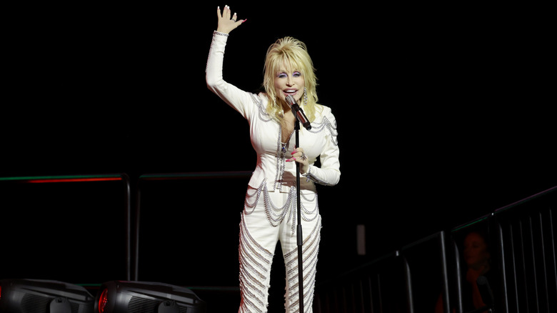 Dolly Parton throws a hand in the air in an all-white outfit with silver detailing on stage