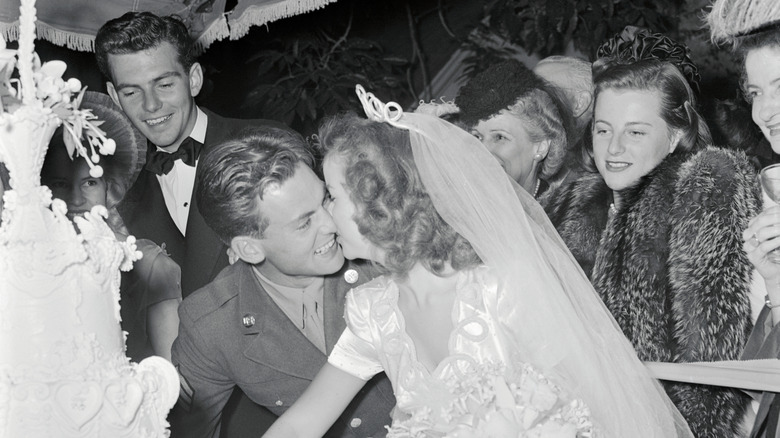 Shirley Temple and John Agar kissing as they cut their wedding cake while guests watch and smile from behind them