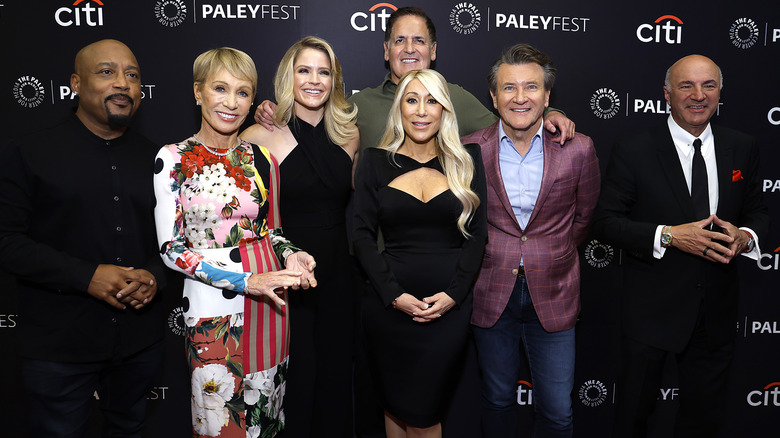 The cast of Shark Tank pose together in front of a black backdrop.