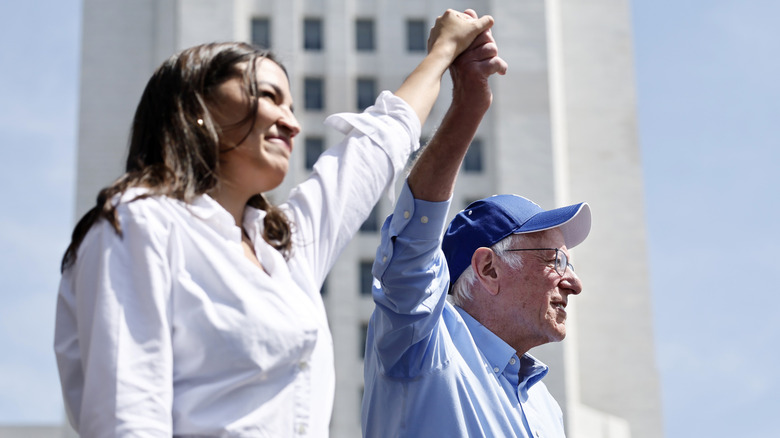 Alexandria Ocasio-Cortez and Bernie Sanders holding hands in the air addressing a crowd on their 'Fight the Oligarchy' tour