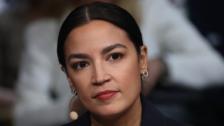Alexandria Ocasio-Cortez wearing a microphone and glancing pensively ahead of her