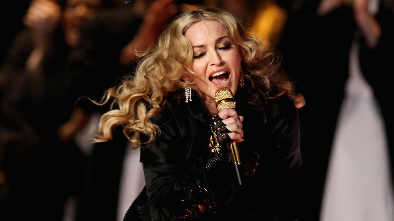 Madonna singing into a microphone at the 2012 Superbowl with long flowing blonde curls blowing in the wind
