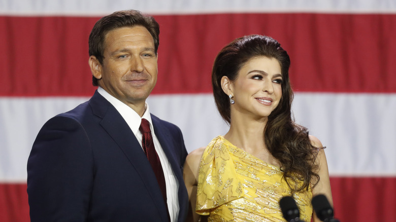 Ron and Casey Desantis smiling at a crowd in front of an American Flag