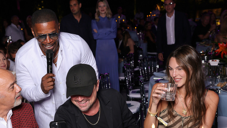 Leonardo DiCaprio smiling, seated next to Vittoria Ceretti while Jamie Foxx stands behind and speaks into a microphone