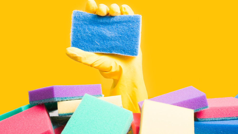 Yellow gloved hand holding a blue sponge amid a pile of colorful sponges