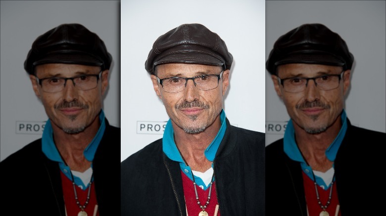Michael Nader poses wearing blue collared shirt, black jacket, glasses, and a leather hat.