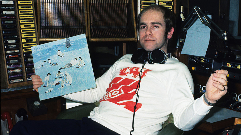 A young Elton John sitting in a studio and holding an album in 1976