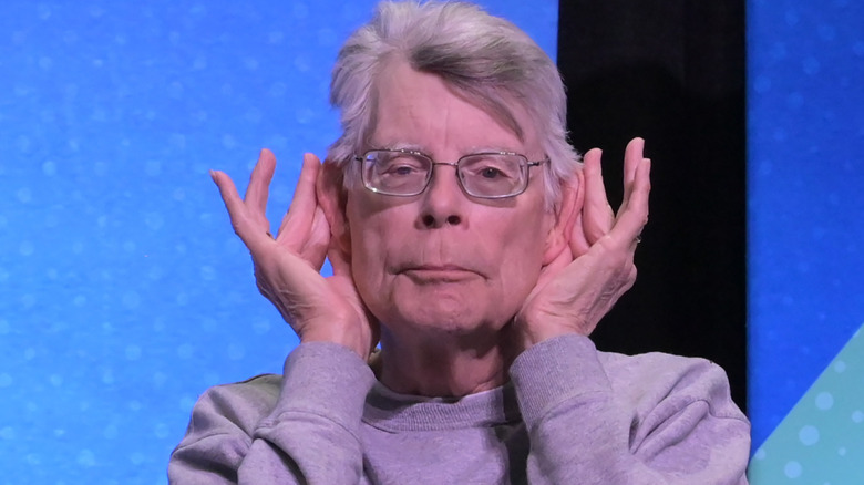 Stephen King holding his ears in a light grey sweater