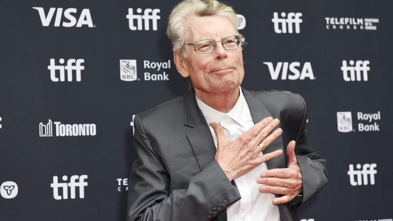 Stephen King placing a hand on his heart while smirking in a grey suit with a white shirt underneath