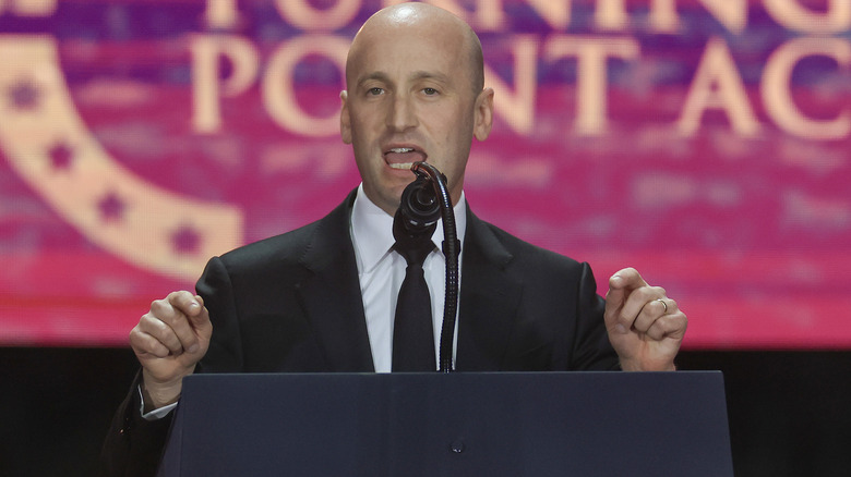 Stephen Miller delivering a speech with his hands raised in a black suit