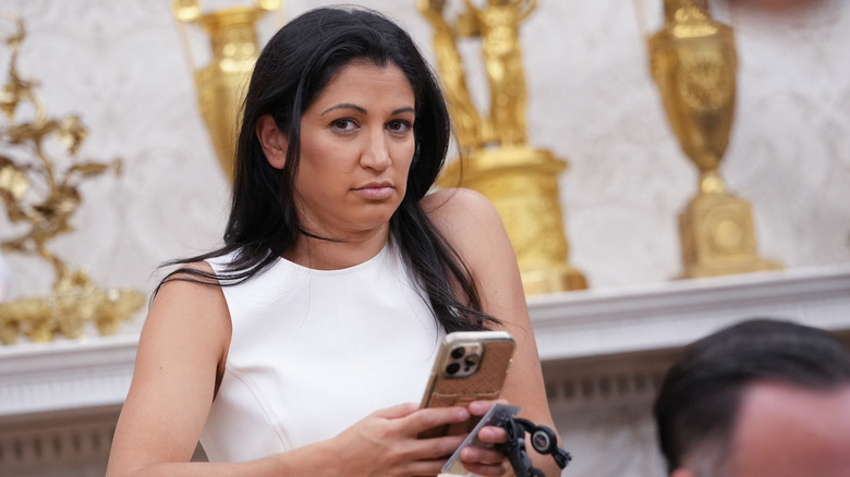 Katie Miller looking at the camera while holding a phone in a white sleeveless dress