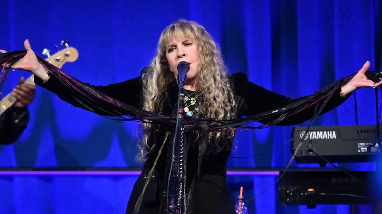 Stevie Nicks singing into a mic while wearing a black dress and stretching her arms out wide.