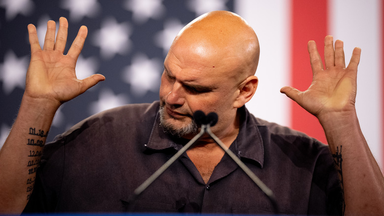 John Fetterman standing in front of an American flag with his hands raised.