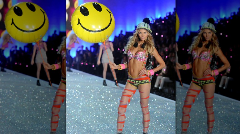 Jessica Hart in cap and headphones wearing colorful lingerie and holding happy face balloon
