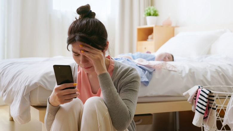 Woman looks at phone while baby is on bed