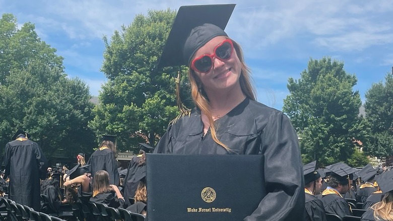 Rowan Henchy smiles in red sunglasses on her college graduation day wearing a black robe holding her diploma