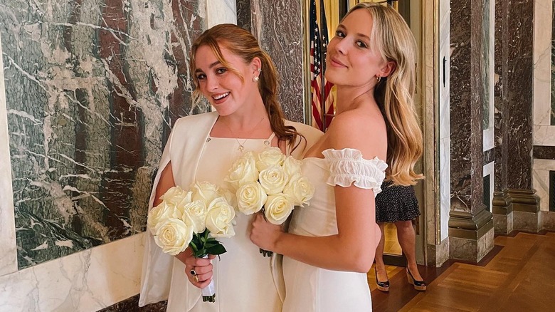 Rowan smiles next to her friend on her high school graduation holding flowers and wearing a white dress