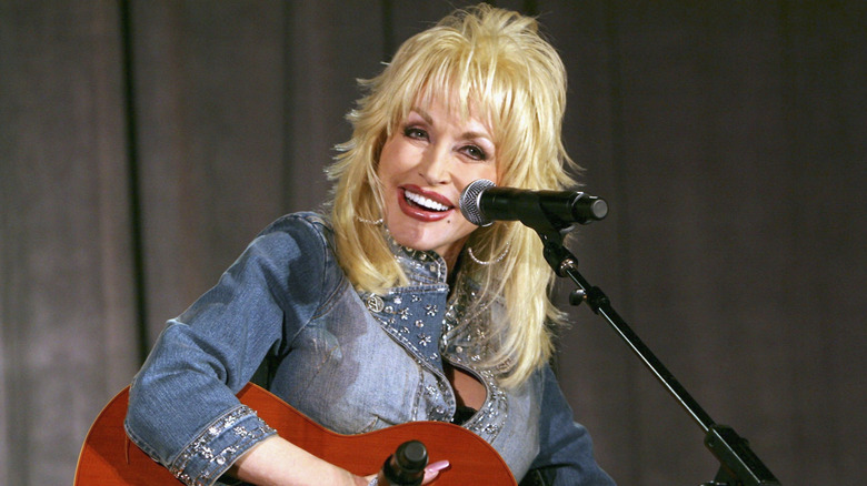 Dolly Parton smiles while playing a guitar and wearing a denim top