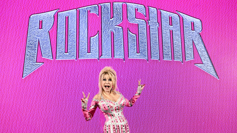Dolly Parton poses with double peace fingers in front of a sign that reads "ROCKSTAR"