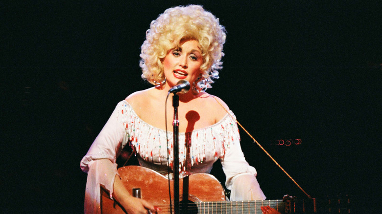 A young Dolly Parton plays an acoustic guitar and sings into a mic