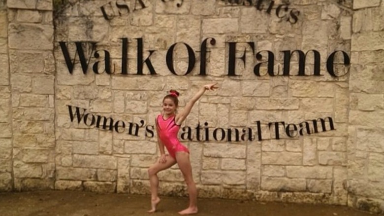 Olivia Dunne as a child posing in front of USA Gymnastics Walk of Fame Women's National Team
