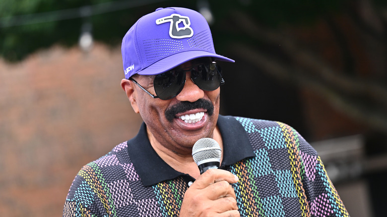 Steve Harvey clutching a microphone and grinning in a colorful polo shirt and baseball hat at a golf game in 2026.