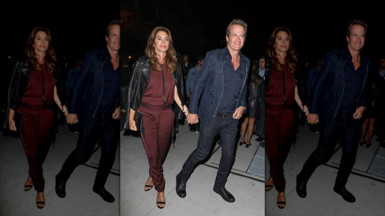 Rande Gerber and Cindy Crawford walking hand and hand at a fashion show, while cindy wears a burgundy jumpsuit with black zippers and a black leather jacket draped over her shoulders