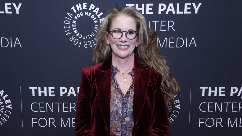 Melissa Gilbert smiling while posing for photos in a velvet blazer, purple patterned blouse, and glasses