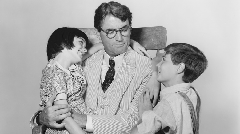 Mary Badham smiling and sitting on Gregory Peck's lap while he looks at Phillip Alford