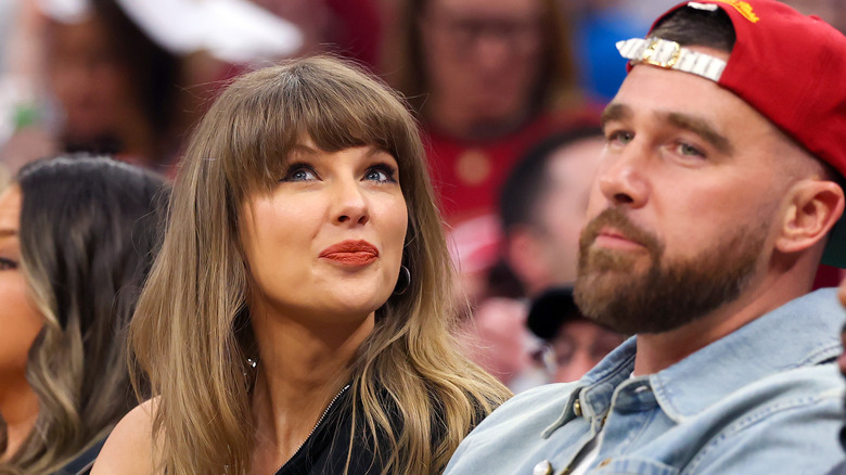 A smiling Taylor Swift looking up while sitting next to Travis Kelce