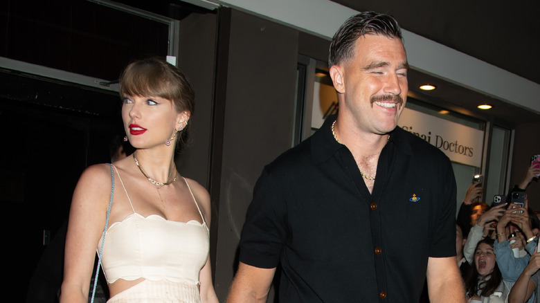Taylor Swift wearing a white cutout dress with a floral pattern holding Travis Kelce's hand as he smiles in a black shirt