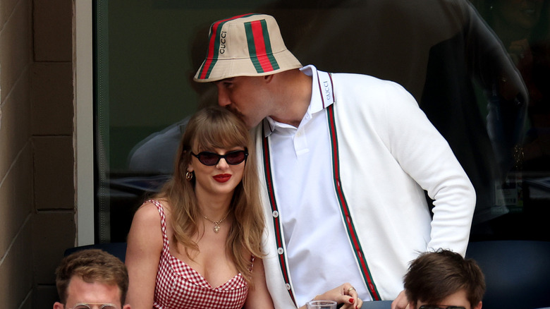 Travis Kelce giving Taylor Swift a kiss on the head in matching white and red outfits and Kelce in all Gucci