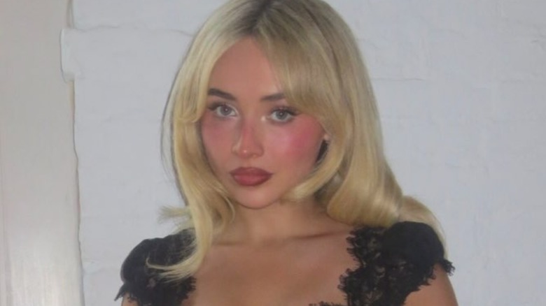 A close-up of Sabrina Carpenter in a black lace dress standing against a white wall