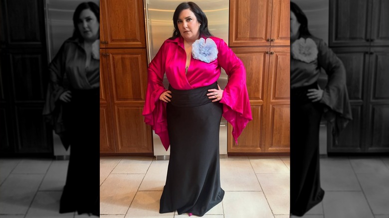 Lena Dunham in a pink blouse and black skirt, with her hands on her hips