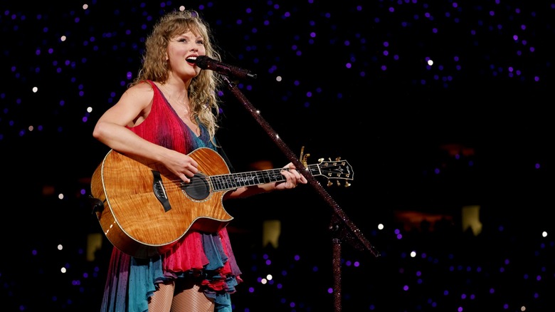 Taylor Swift in a multi-colored dress playing the guitar on stage