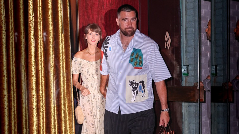 Taylor Swift in a cream peasant dress with Travis Kelce in a blue striped shirt leaving an event