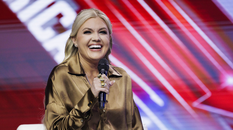 Erika Kirk wearing an olive top and laughing while holding a microphone.