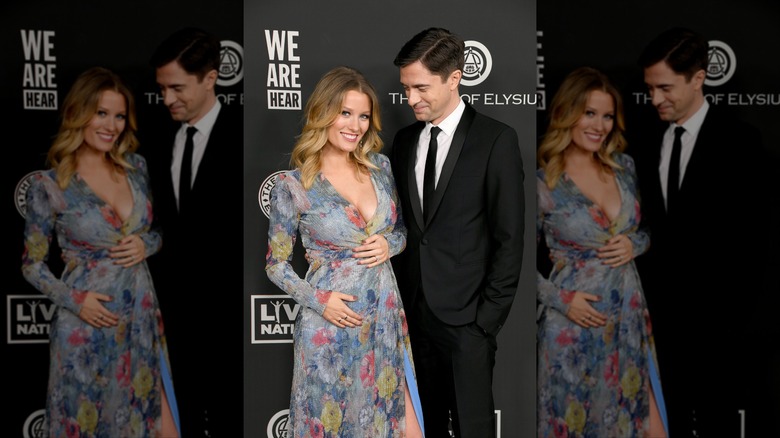 Topher Grace smiling proudly at his wife, Ashley, holding her baby bump on the red carpet