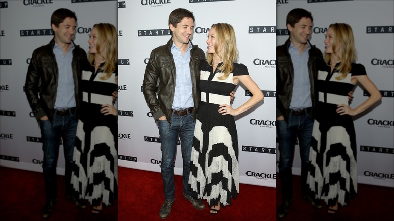 Topher Grace and his wife Ashley smiling at each other while posing for cameras on the red carpet