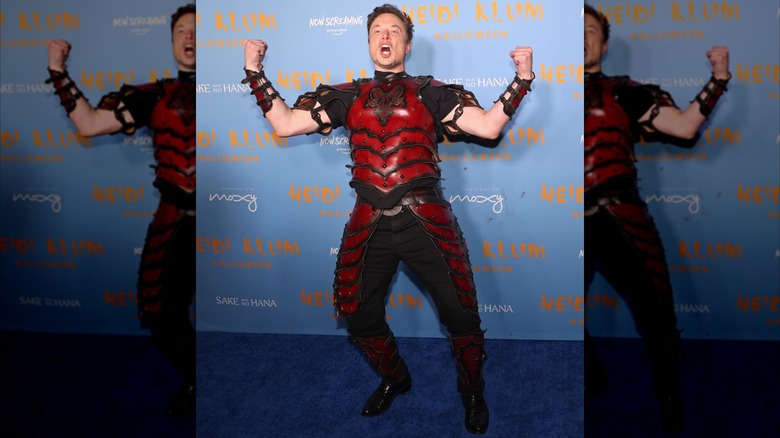 Elon Musk wearing red armor emblazoned with Baphomet head and raising fists in the air at Heidi Klum's 2022 Halloween party