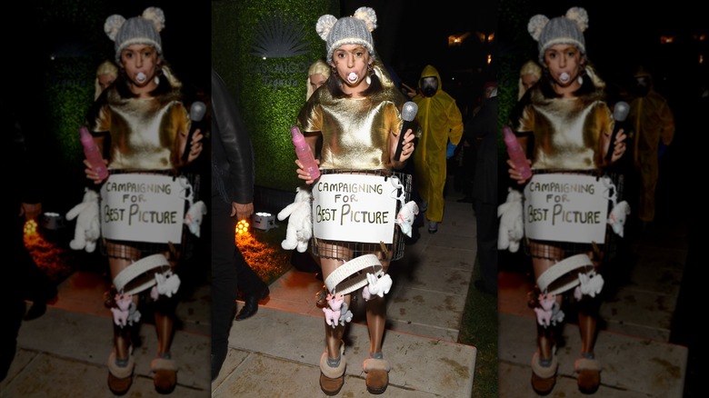 Nina Dobrev wearing her "A Star is Born" costume, including a pacifier, baby bottle, microphone, toys, hat with mouse ears, and "Campaigning for Best Picture" sign