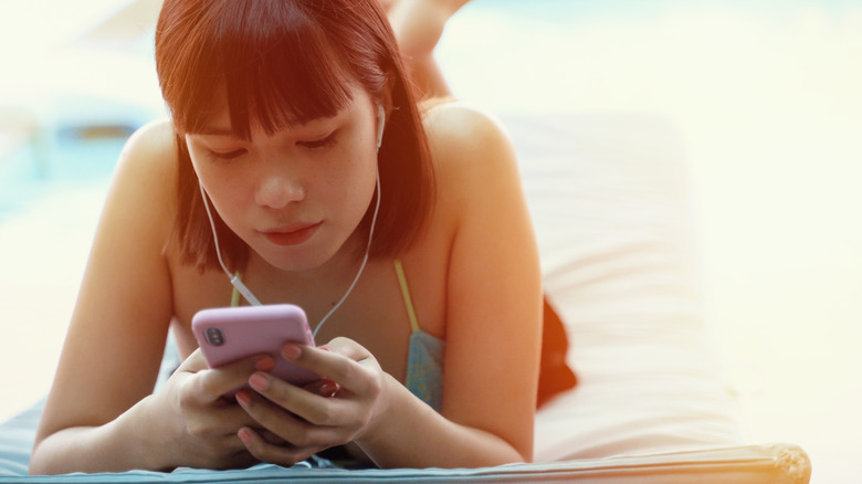 Asian woman laying on stomach on beach tall listening to smartphone with earphones