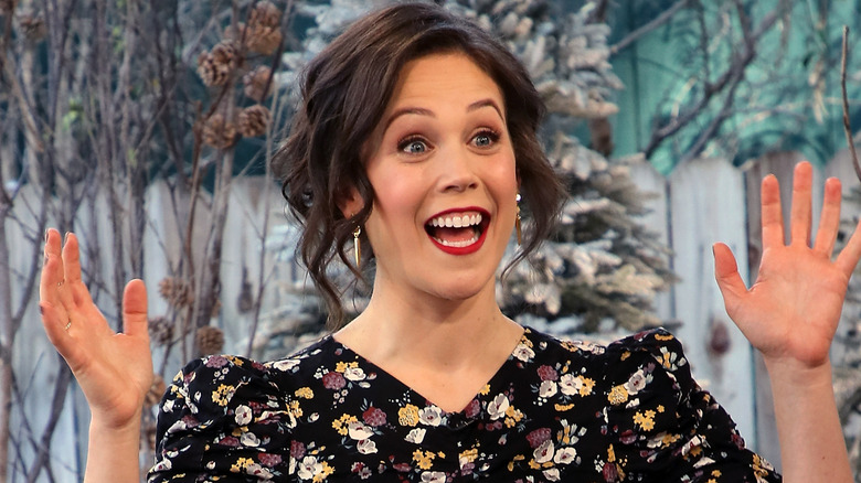 Erin Krakow smiling and holding her hands up in surprise in a floral dress and red lipstick