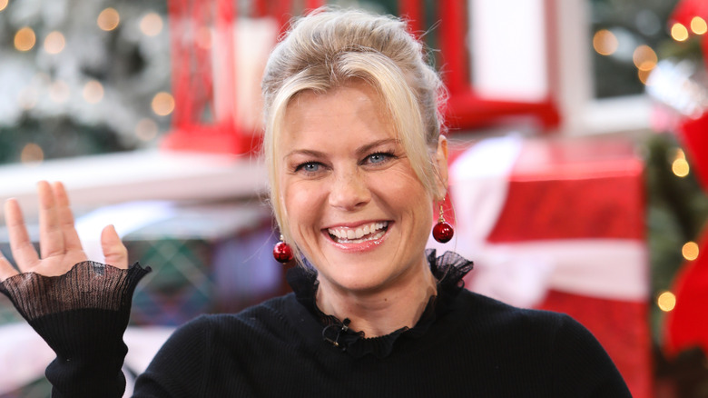 Alison Sweeney smiling and waving in a black long-sleeved dress and festive earrings
