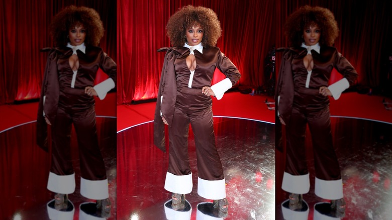 Fleur East posing in a shiny brown jumpsuit with large white cuffs on the sleeves and legs.