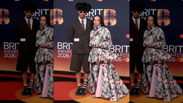 Jordan Stephens wearing a brown dress coat, tie, and shorts while posing with Jade Thirwall, who is wearing a puffy floral-print coat and matching floor-length skirt.