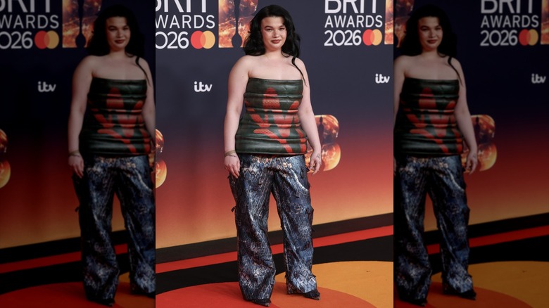 Lola Young posing in a black and red tube top and dark baggy pants with a mesh pattern and metallic details.