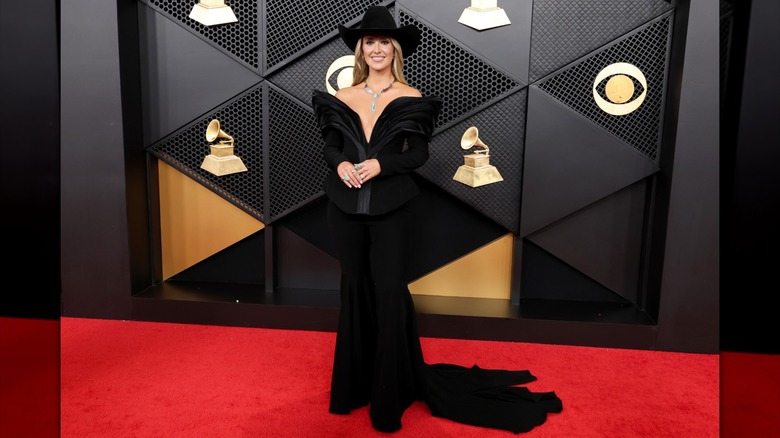 Lainey Wilson smiling in a black cowboy hat and matching off-the-shoulder dress with a train