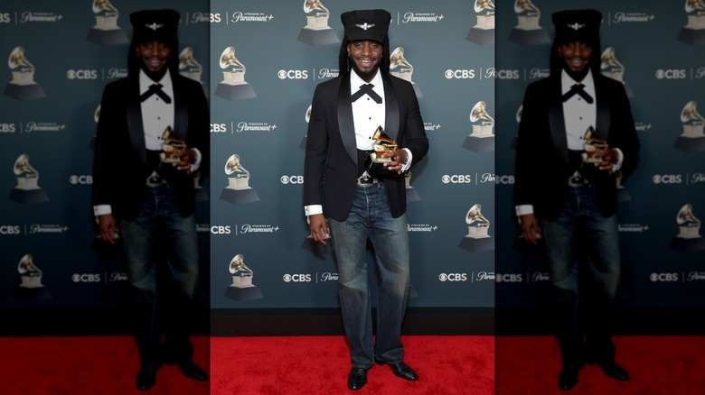 Shaboozey smiling in a black conductor-style hat, matching coat, and blue jeans while holding a Grammy